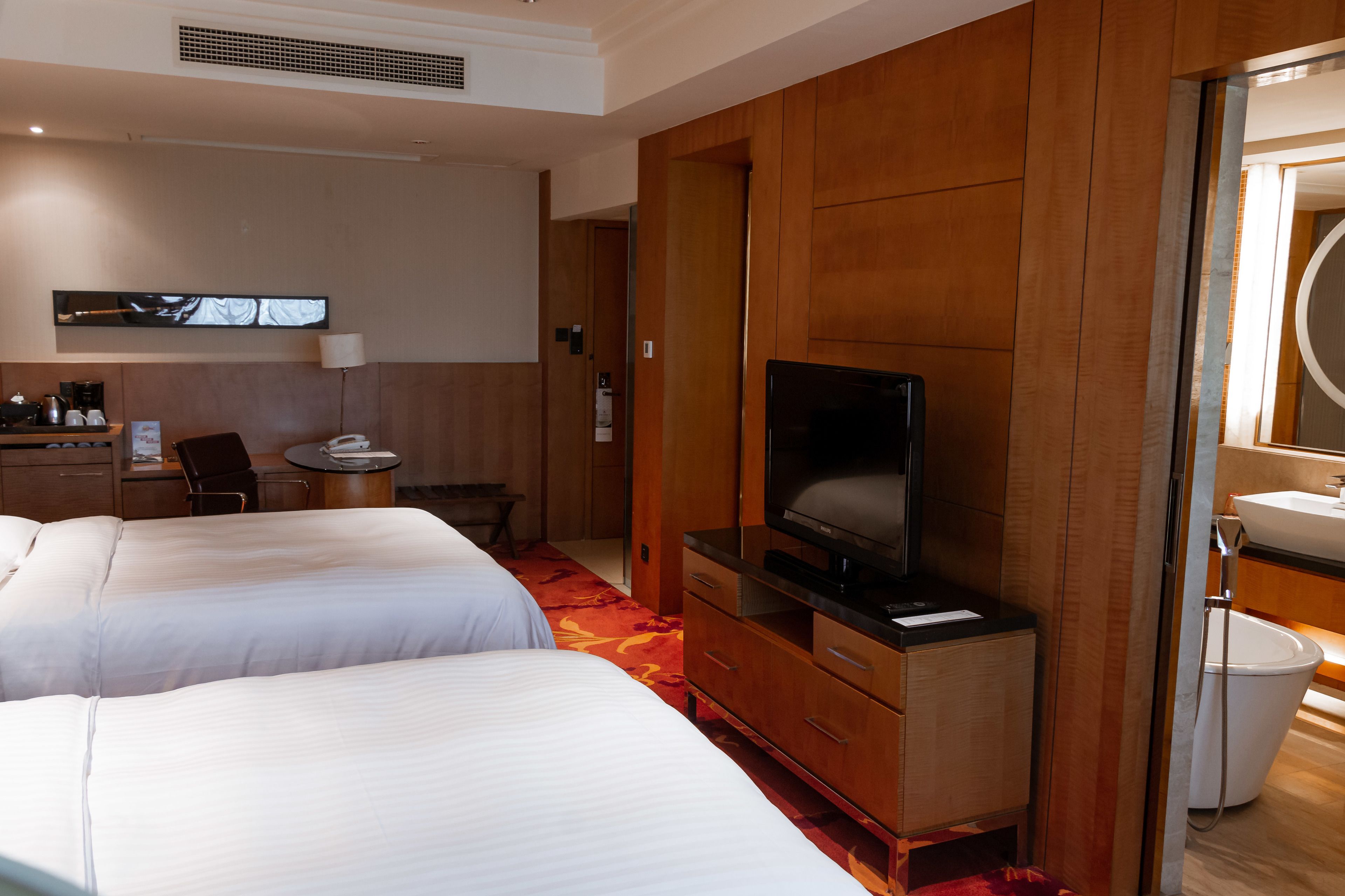 A hotel room with two beds, a TV, a desk with a chair, a desk lamp, a coffee maker, and a bathroom with a sink and toilet.
