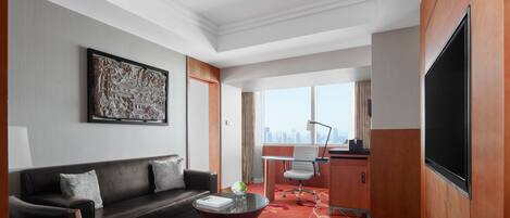 A modern hotel room with a sofa, a glass coffee table, a desk, and a large TV.