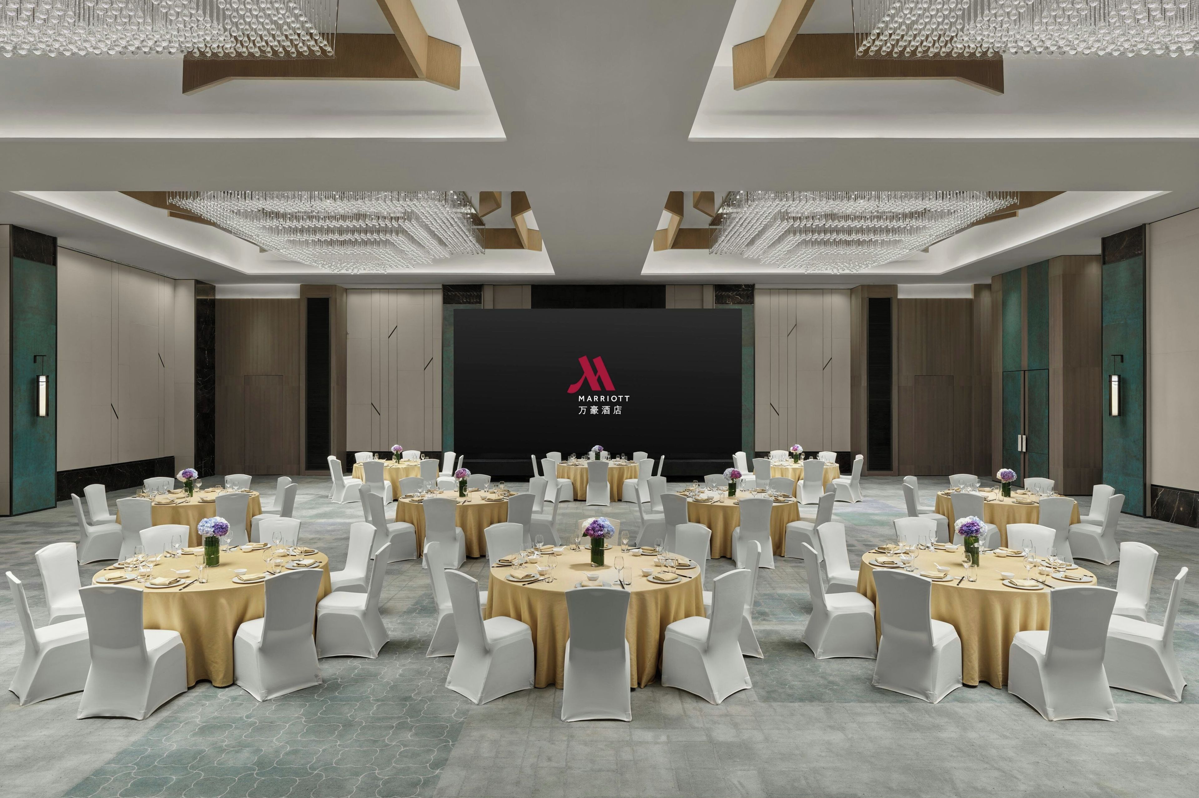 Un salón de conferencias con varias mesas redondas, sillas blancas y una pantalla grande que muestra un logotipo.