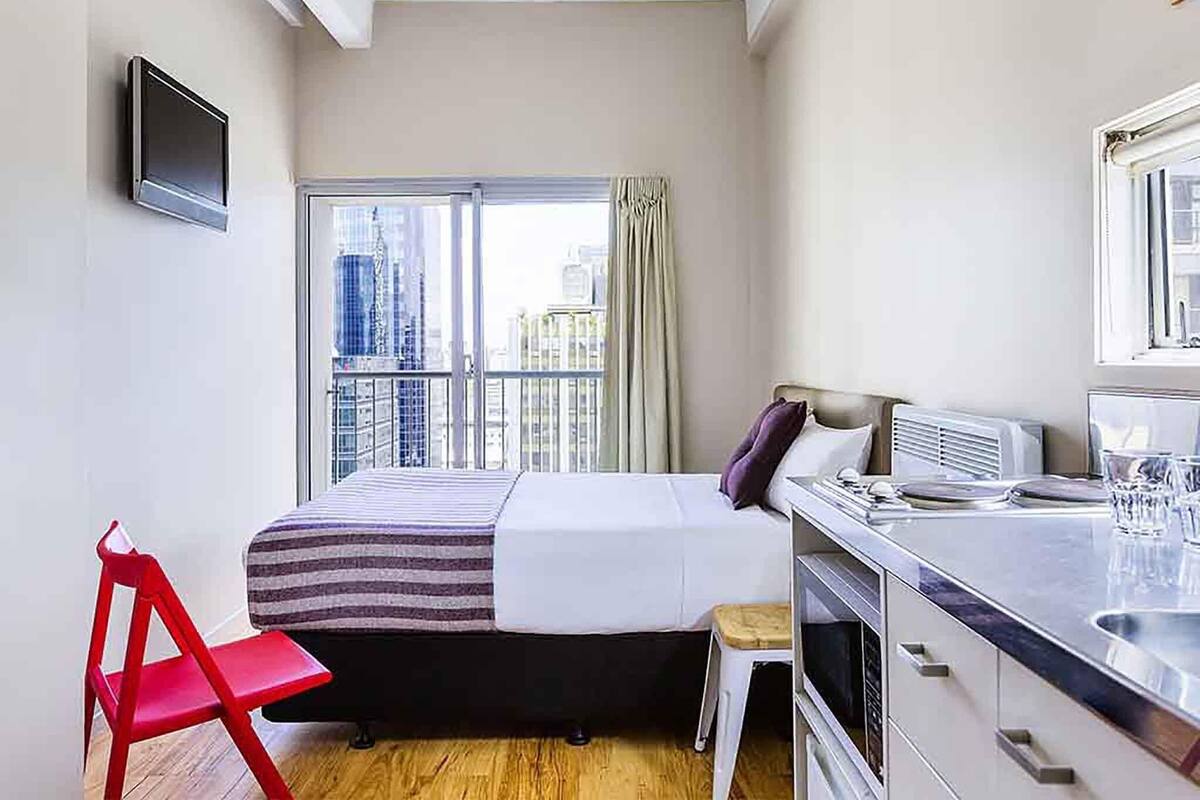 A bedroom with a bed, a red chair, a small stool, a TV, and a window with a city view.