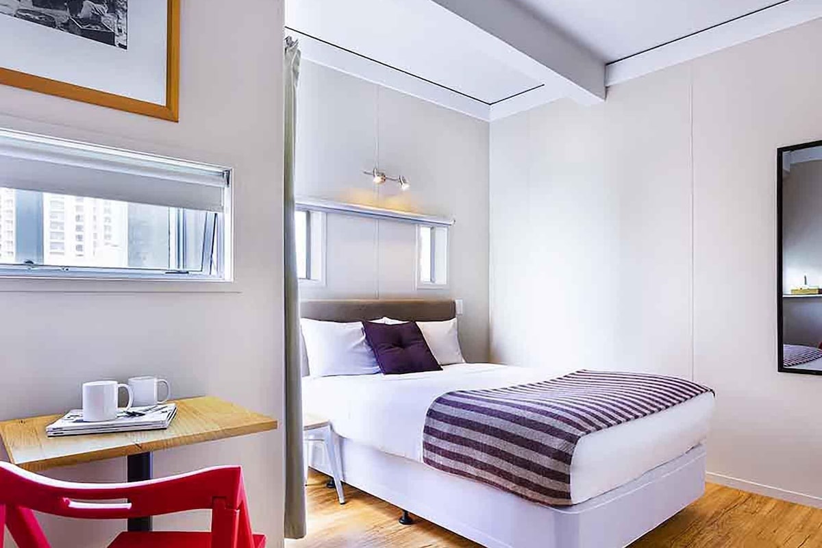 Hotellihuone, jossa on sänky, työpöytä tuolilla, ikkuna ja seinälle asennettu televisio.