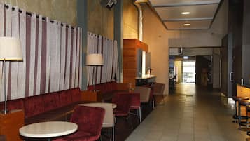 Un hall d’hôtel moderne avec un long comptoir d’accueil, un espace salon avec des fauteuils rouges et un sol carrelé.