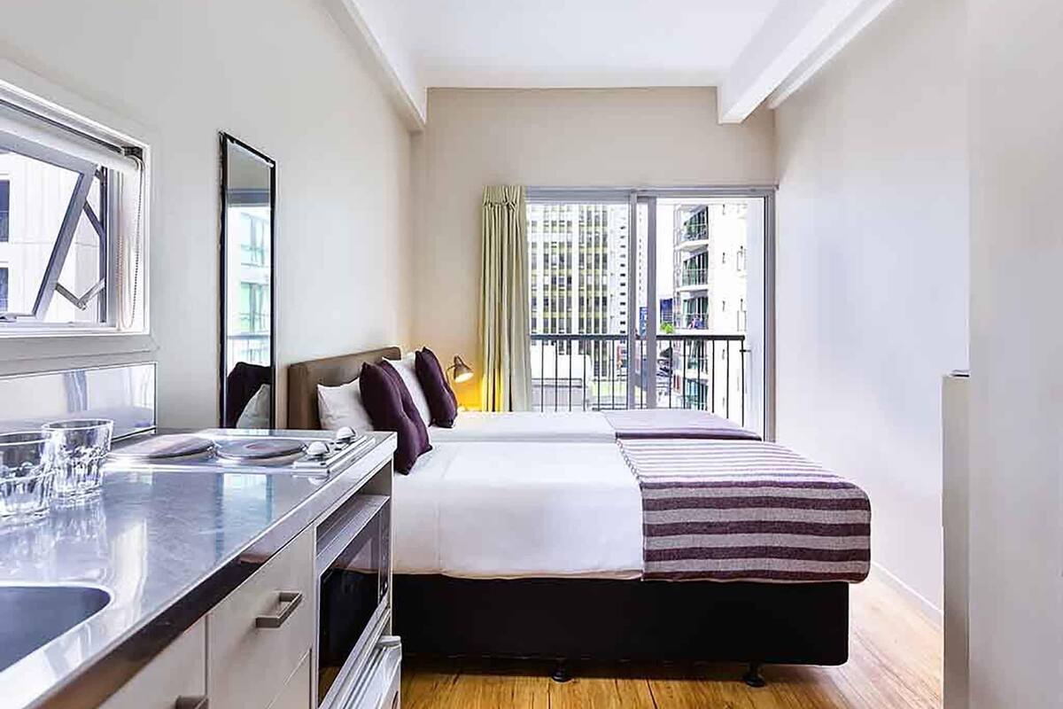 A modern hotel room with a bed, a small kitchenette, and a balcony with a city view.