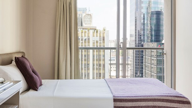Una camera d'albergo con un letto, un comodino e una finestra vista su alti edifici.