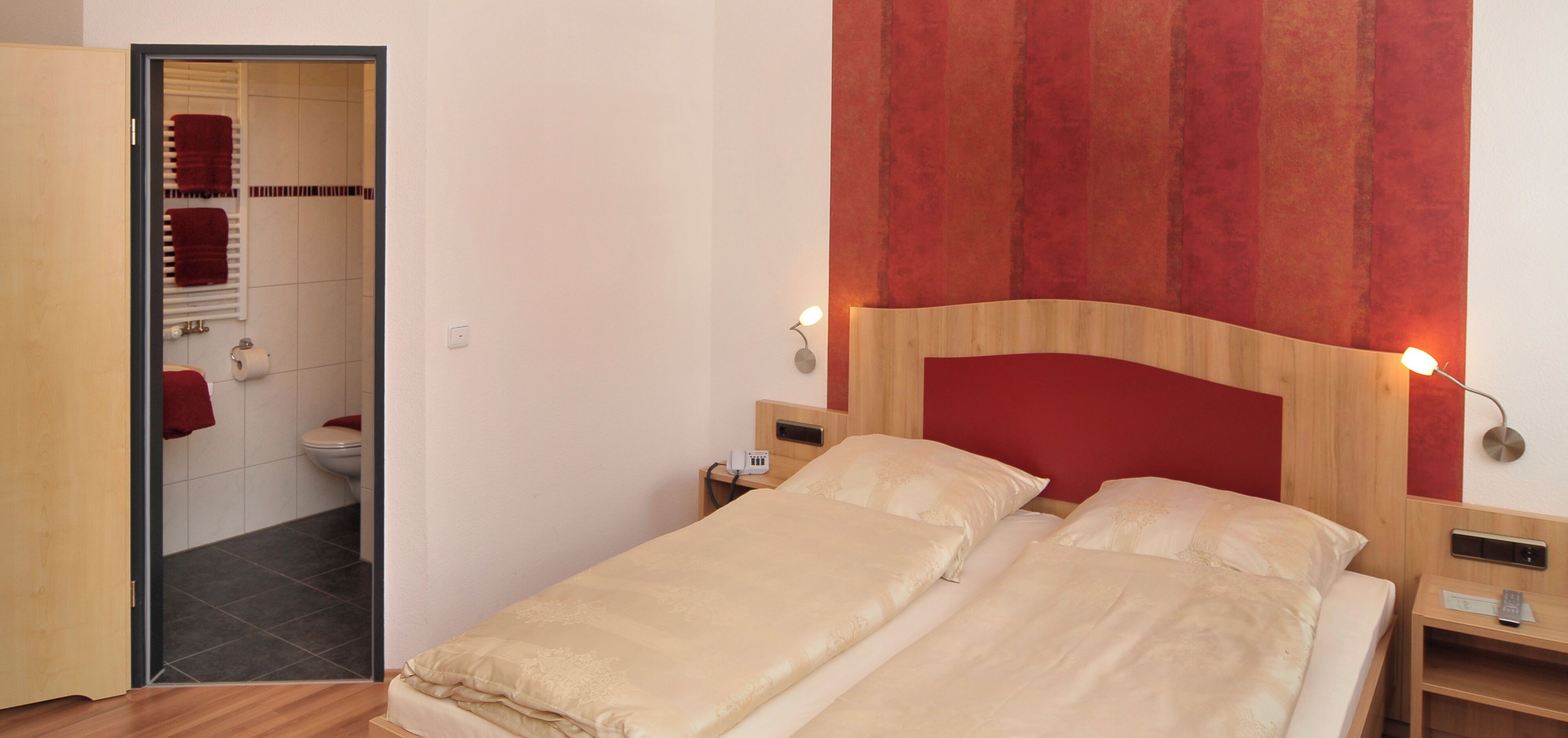 A hotel room with a bed, bedside tables, a nightstand, a red curtain, a wall-mounted lamp, and a bathroom visible through an open door.