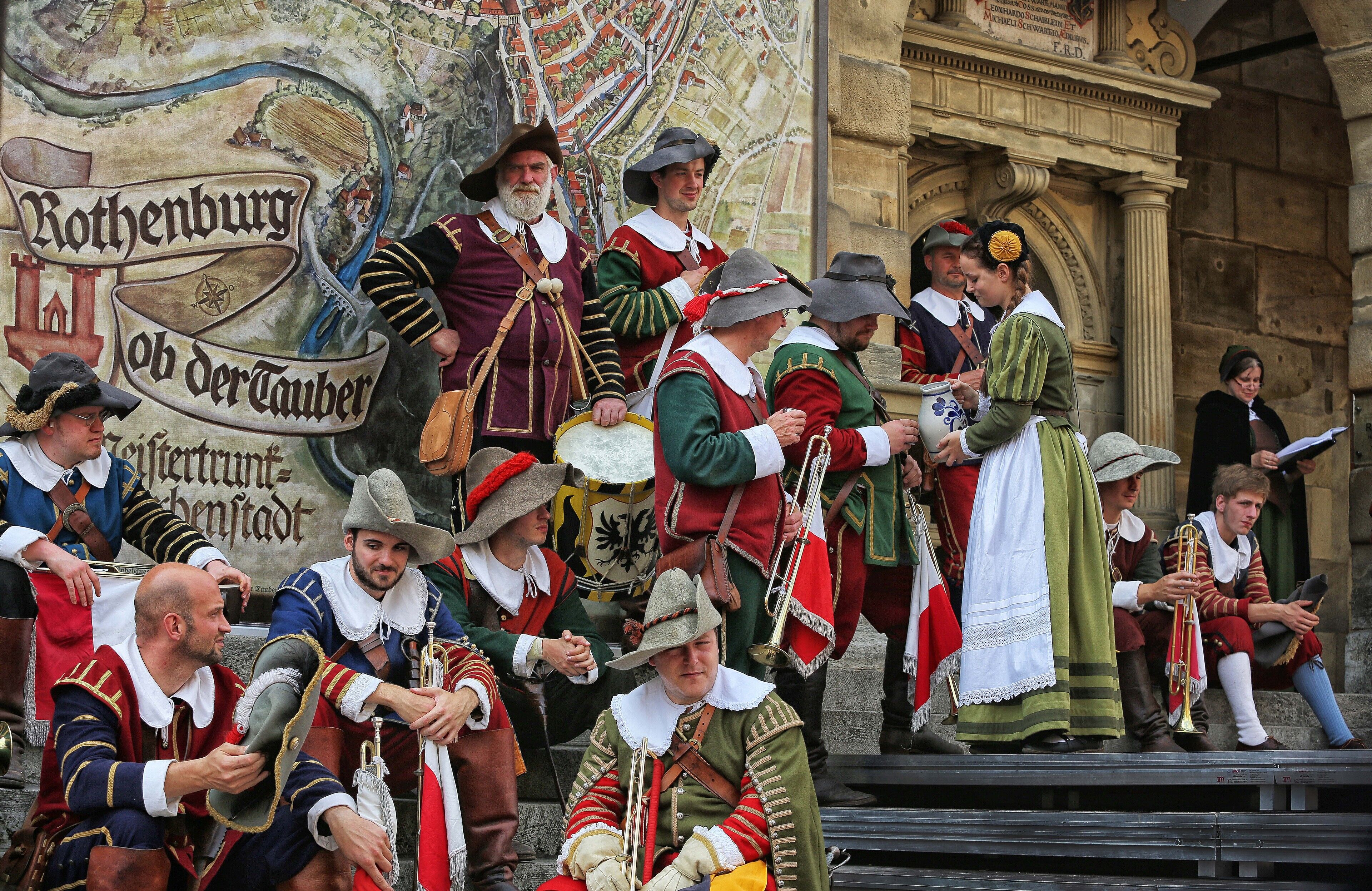Un grupo de personas vestidas con trajes de época, quizás en una fiesta o recreación histórica, posando frente a un edificio con un gran mural.