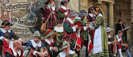 A group of people in historical costumes, possibly at a festival or reenactment, standing in front of a building with a large mural.