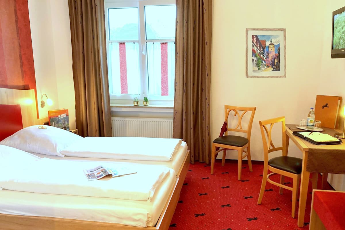 Ein Hotelzimmer mit zwei Einzelbetten, einem Schreibtisch mit Stuhl, einem Fenster mit Vorhängen, einer Heizung und einem gerahmten Bild an der Wand.