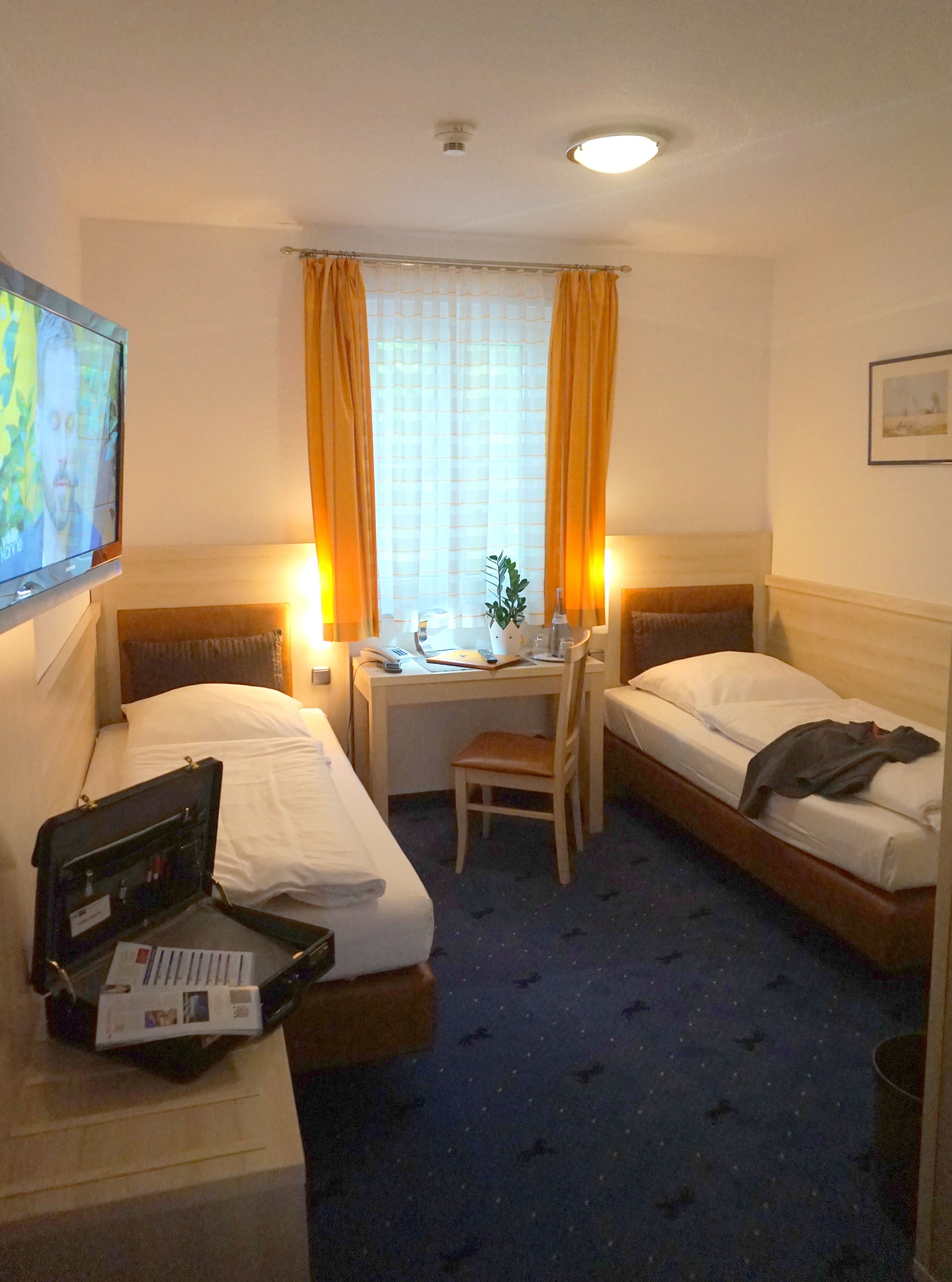A hotel room with two single beds, a desk with a chair, a TV, and a suitcase on the floor.