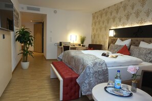 Une chambre d’hôtel avec un grand lit, une table de chevet avec une bouteille d’eau, un plateau avec des verres, une plante en pot et un banc rouge.