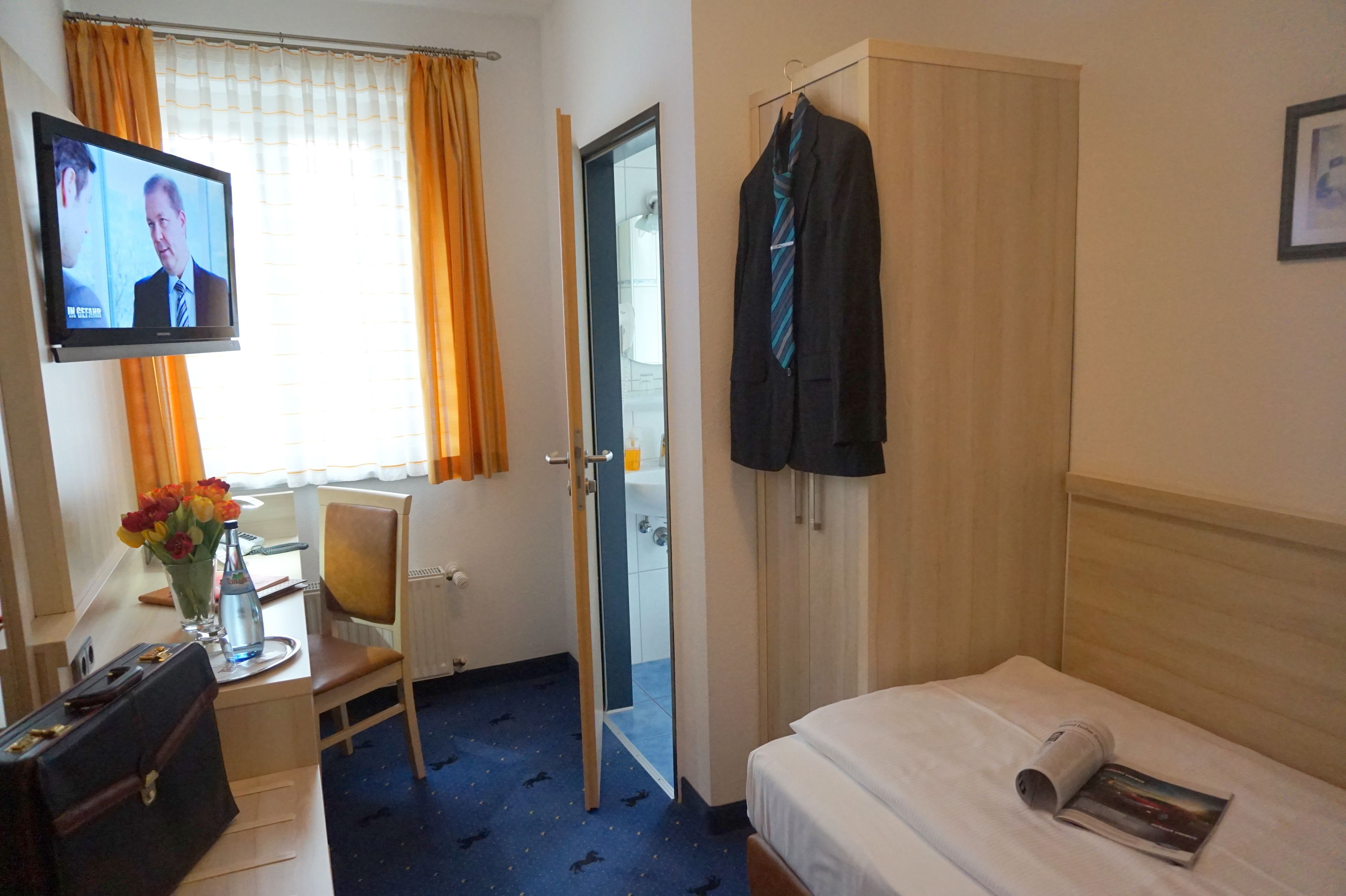 A hotel room with a bed, a desk with a chair, a television, a wardrobe, and a door leading to a bathroom.
