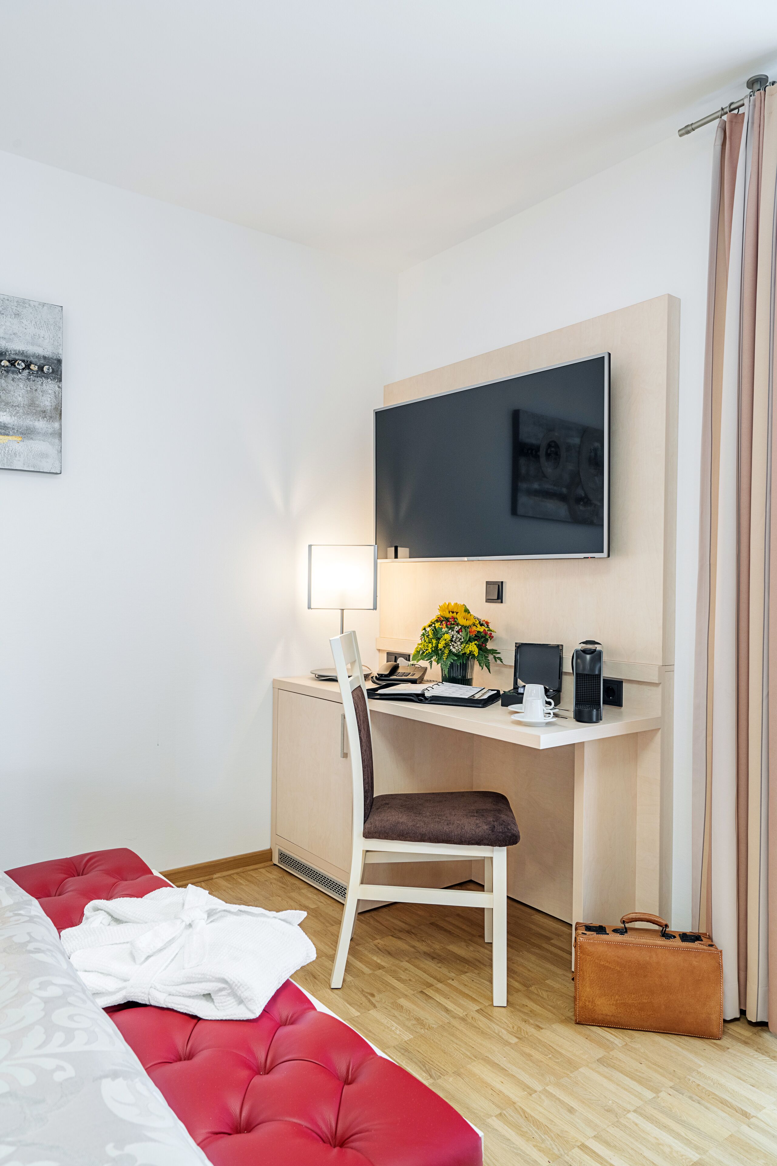 Ein Hotelzimmer mit einem Bett, einem Schreibtisch mit Stuhl, einem Fernseher, einer Lampe, einer Blumenvase und einem Koffer.