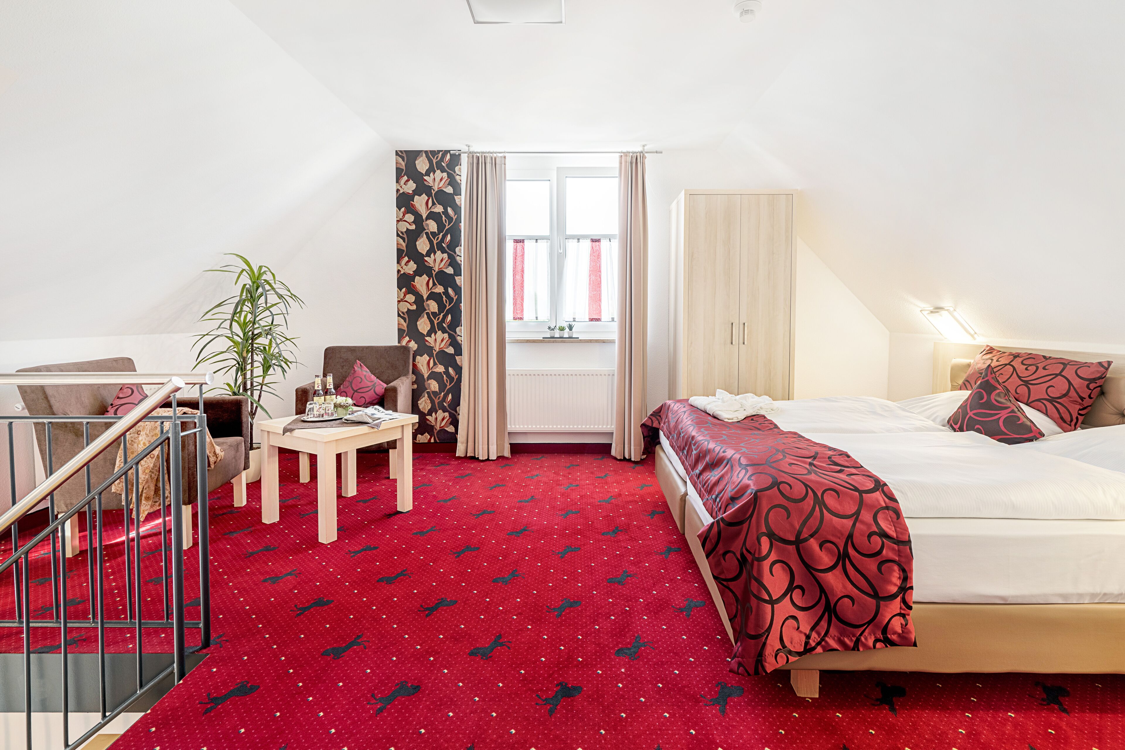 Une chambre d’hôtel avec un lit, un bureau, une chaise, une plante et une fenêtre avec des rideaux.