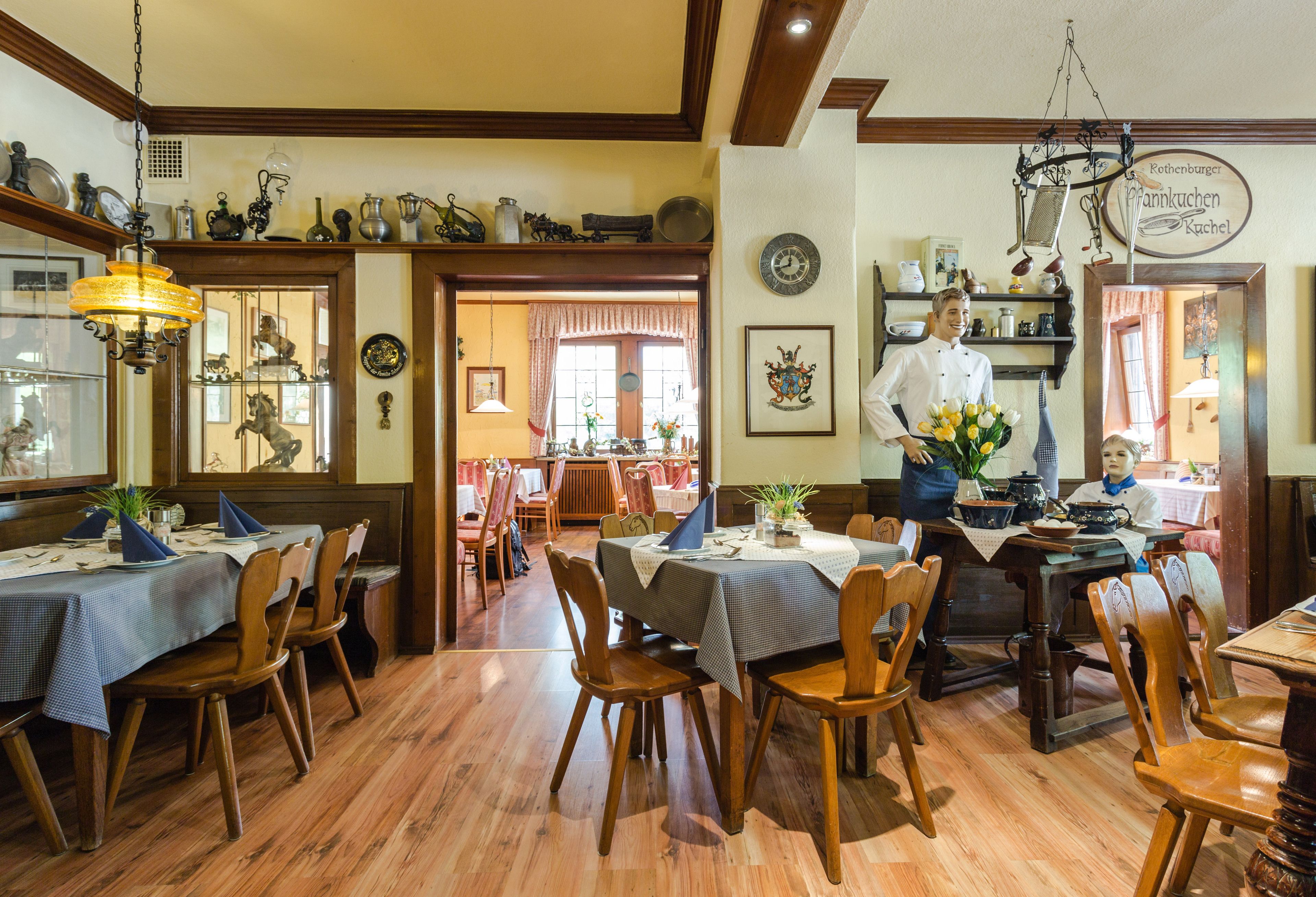 Un restaurante con mesas y sillas de madera, un cocinero al mostrador y diversos utensilios de cocina a la vista.