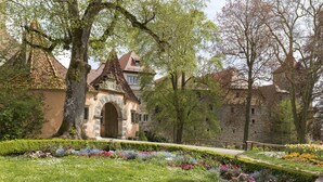 Ein historisches Gebäude mit Bogenportal, umgeben von einem gepflegten Garten mit verschiedenen Blumen und einem Weg dorthin.