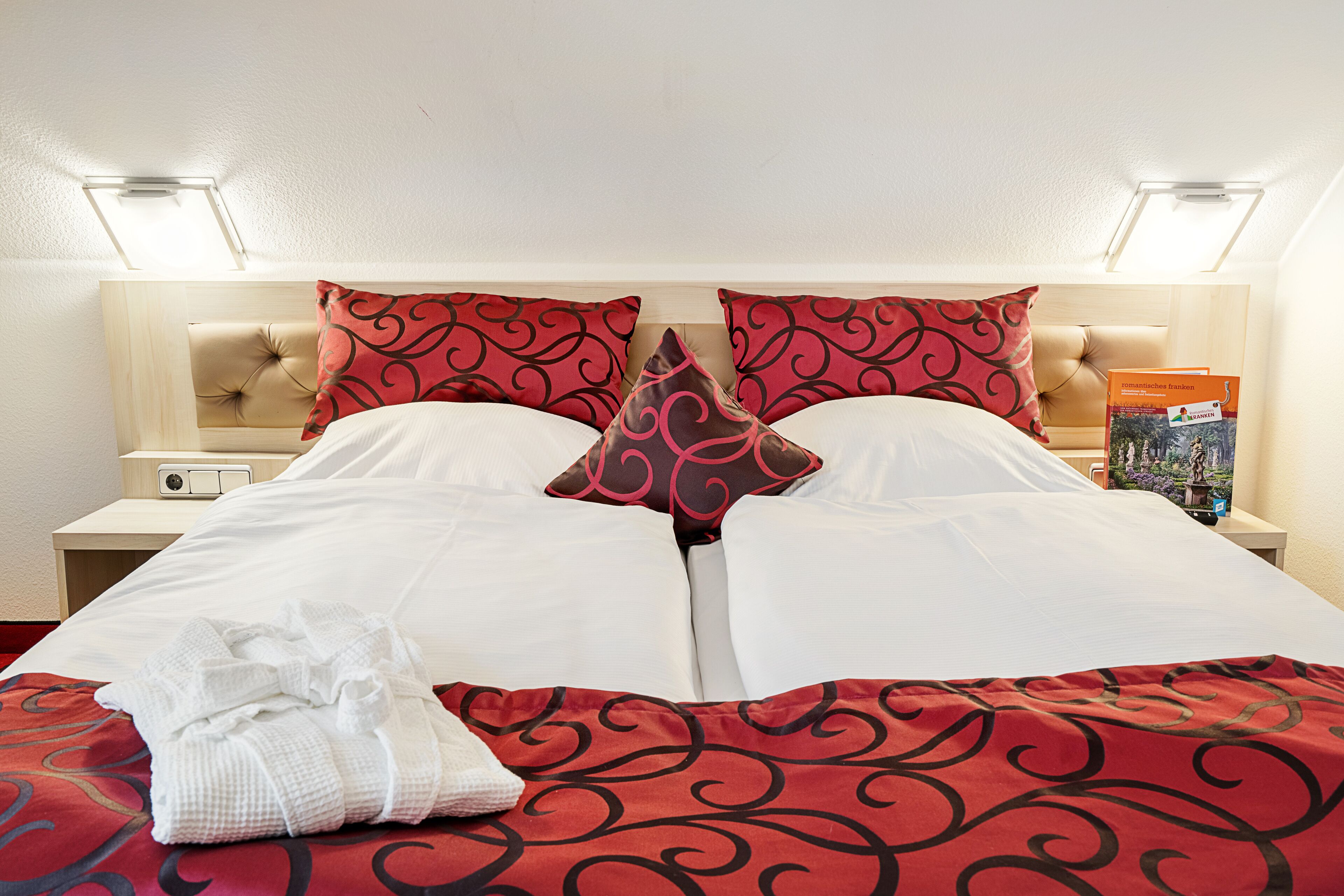 Una habitación de hotel con una cama, dos almohadas, una mesita de noche, una lámpara y un libro sobre la mesita.