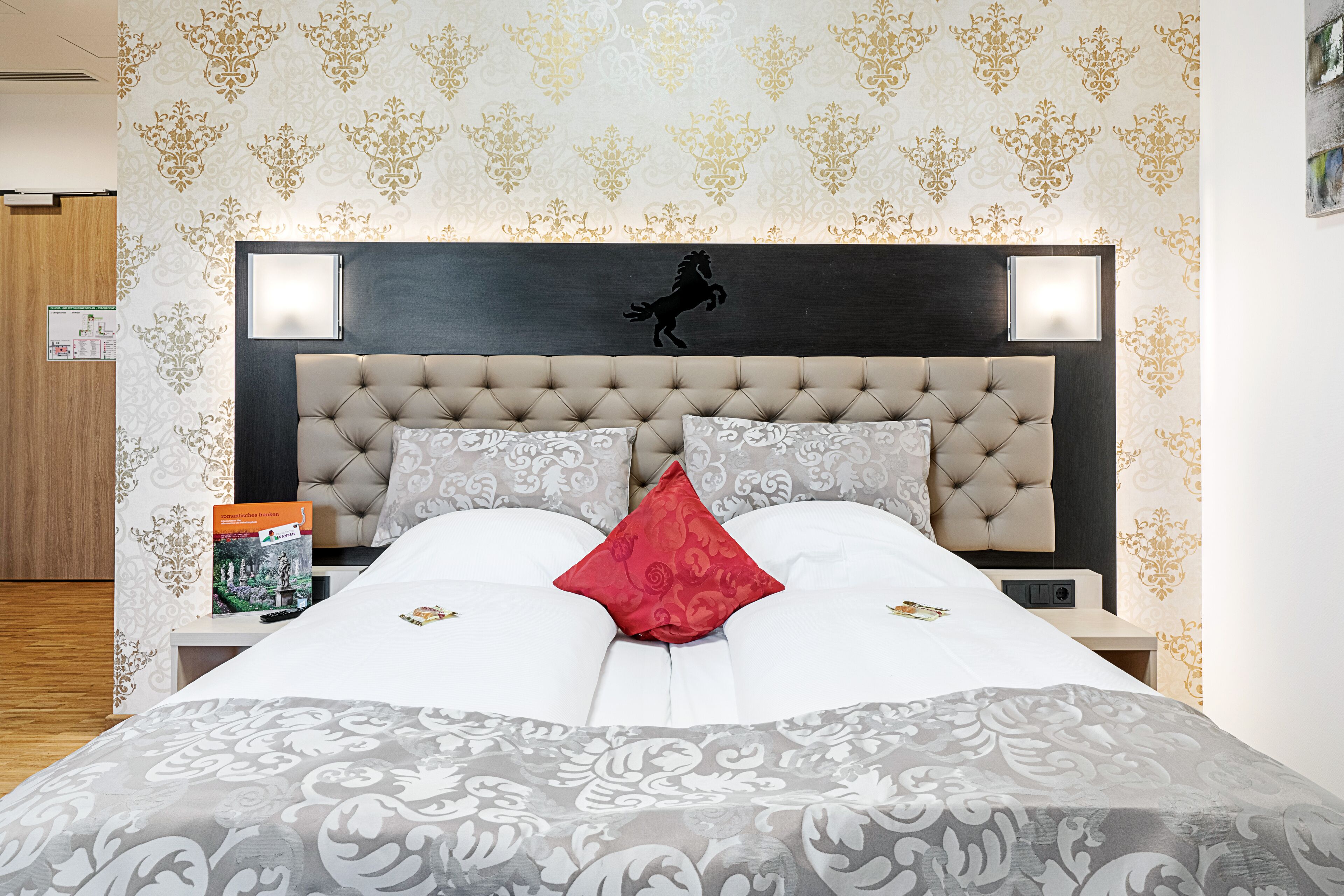 Ein Hotelzimmer mit einem großen Bett, einem Kopfteil mit Pferde-Silhouette, strukturierten Tapeten und einem Nachttisch mit Buch und Fernbedienung.