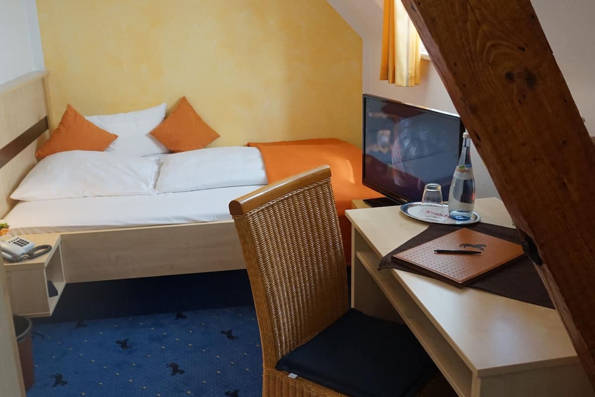 Ein kleiner, gemütlicher Raum mit Bett, Schreibtisch und Stuhl. Ein Fenster lässt natürliches Licht herein, eine Deckenleuchte ist sichtbar.
