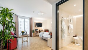 Ein modernes Hotelzimmer mit Holzboden, einem Bett, einem Schreibtisch mit Stuhl, einem Fernseher, einer Pflanze und einem roten Koffer.
