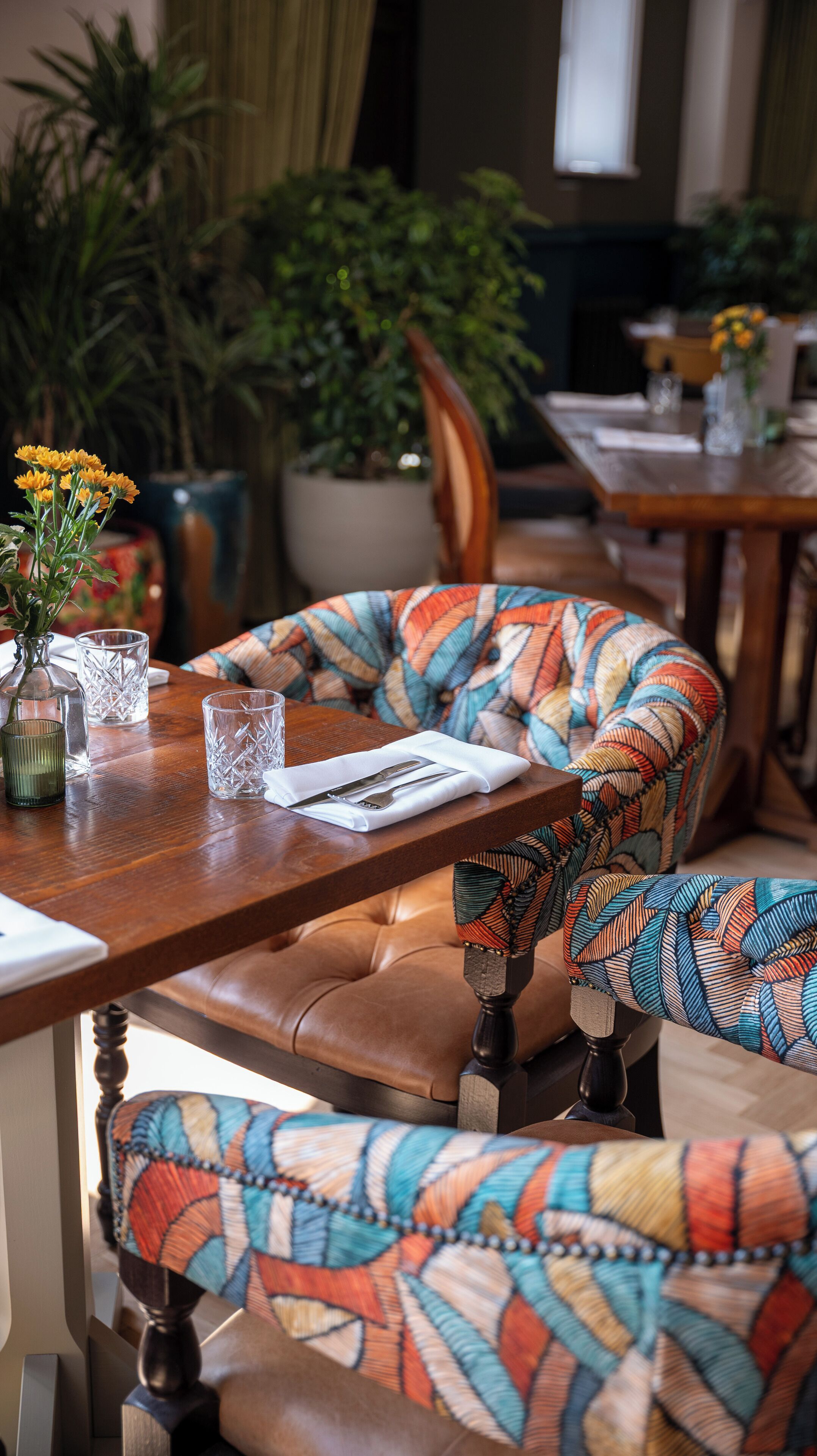 A dining area with a wooden table set for two, featuring a patterned cushion, clear glassware, and a vase with yellow flowers.