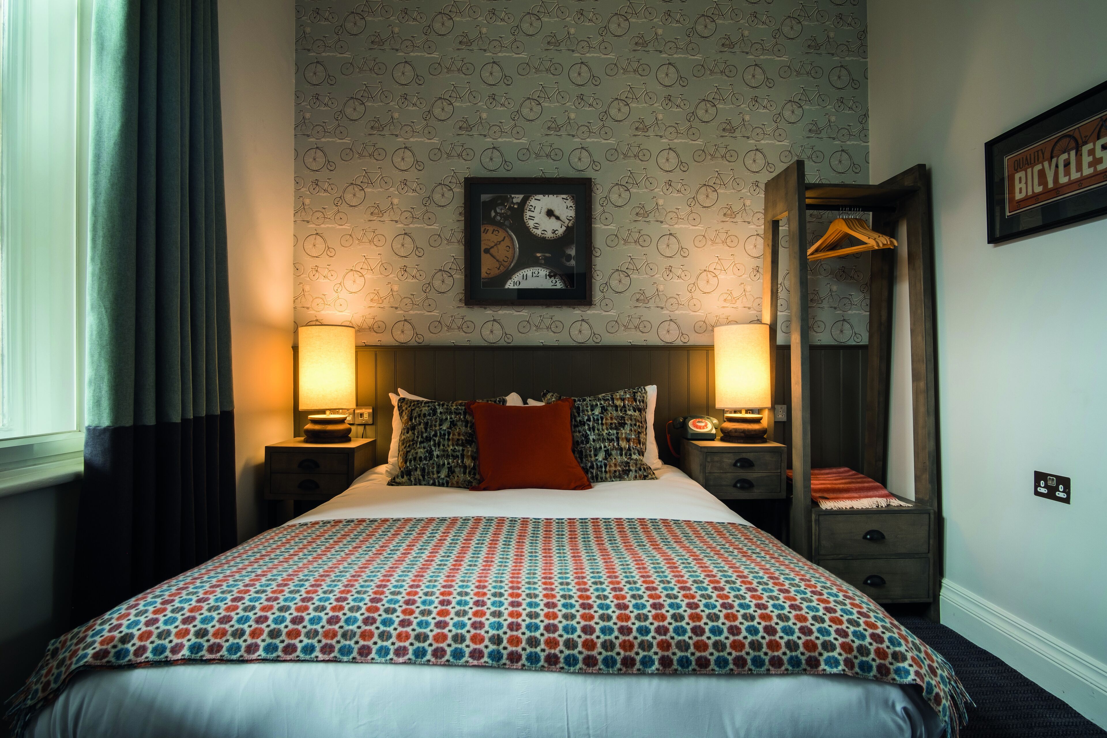 A neatly made bed with patterned bedding, two bedside lamps, a framed picture on the wall, and a bicycle-themed wallpaper.