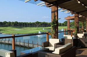 Un espace extérieur aménagé, abrité et doté de mobilier en osier, offrant une vue sur un terrain de golf et un plan d’eau.