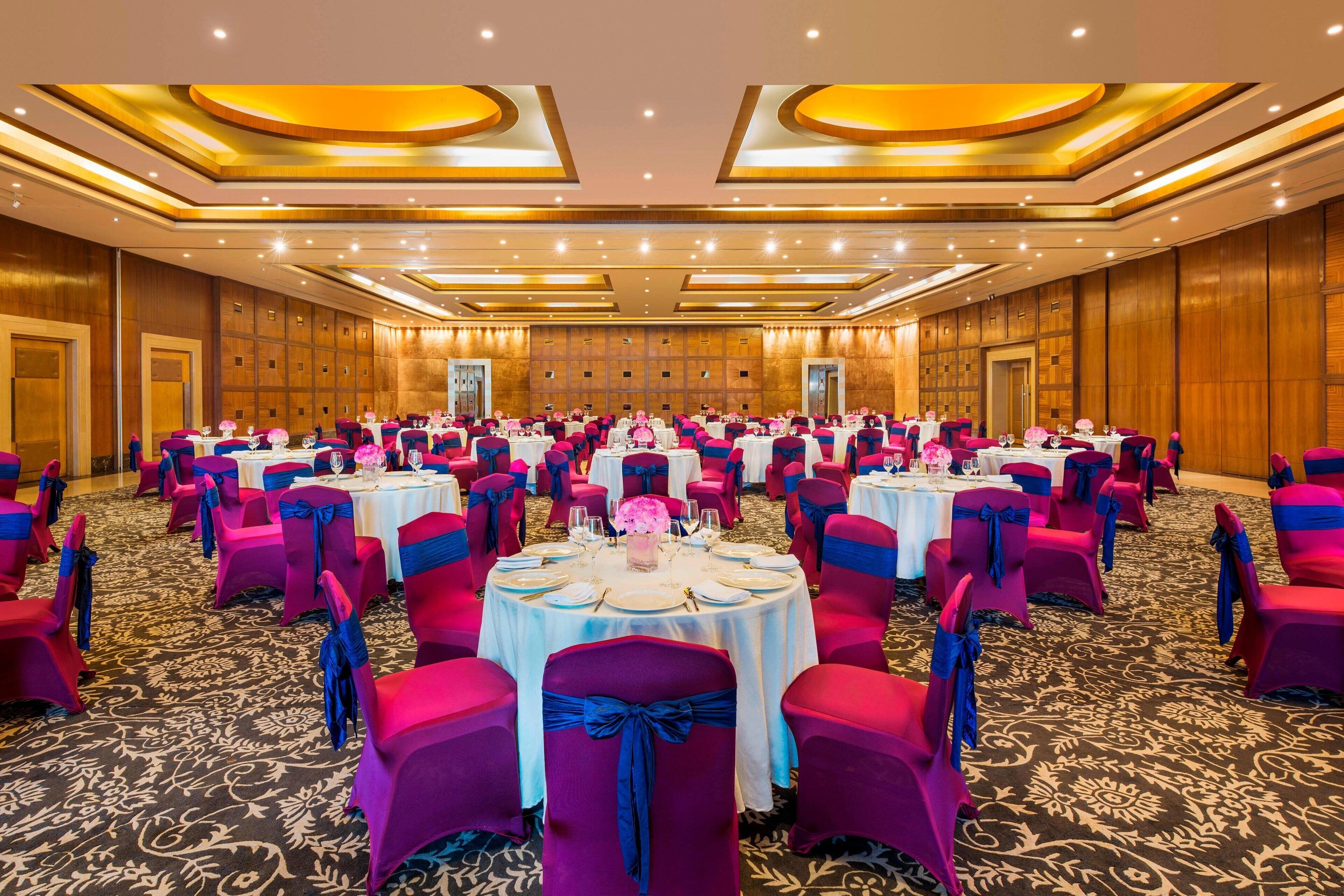 Un gran salón de banquetes con mesas redondas, sillas rosadas y azules, y suelo alfombrado.