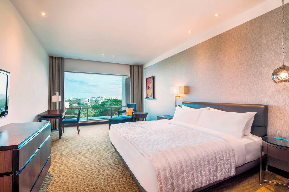 A hotel room with a large bed, a desk, a chair, a TV, and a view of the city.