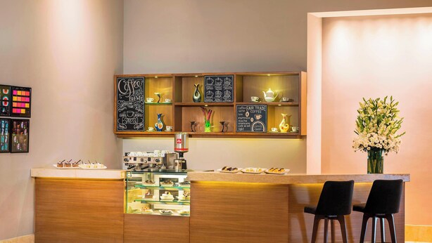 A modern bar area with a wooden counter, a coffee machine, and a display of pastries. There are two black bar stools and a vase of flowers on the right.