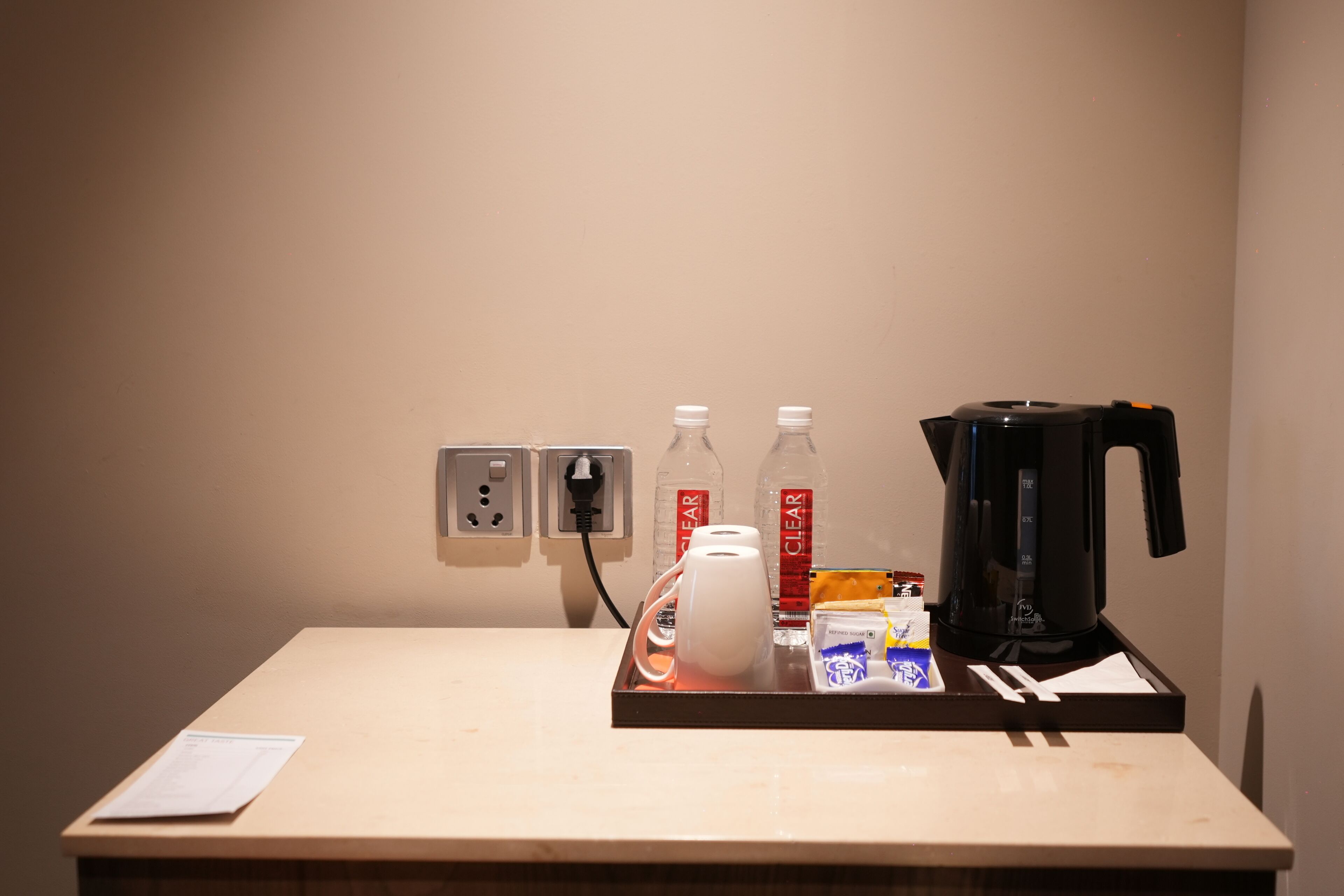 Ein Hotelzimmer mit einem Holztisch, einer weißen Karaffe, zwei klaren Wasserflaschen, einer schwarzen Kaffeemaschine und einer kleinen Tasche Taschentücher.