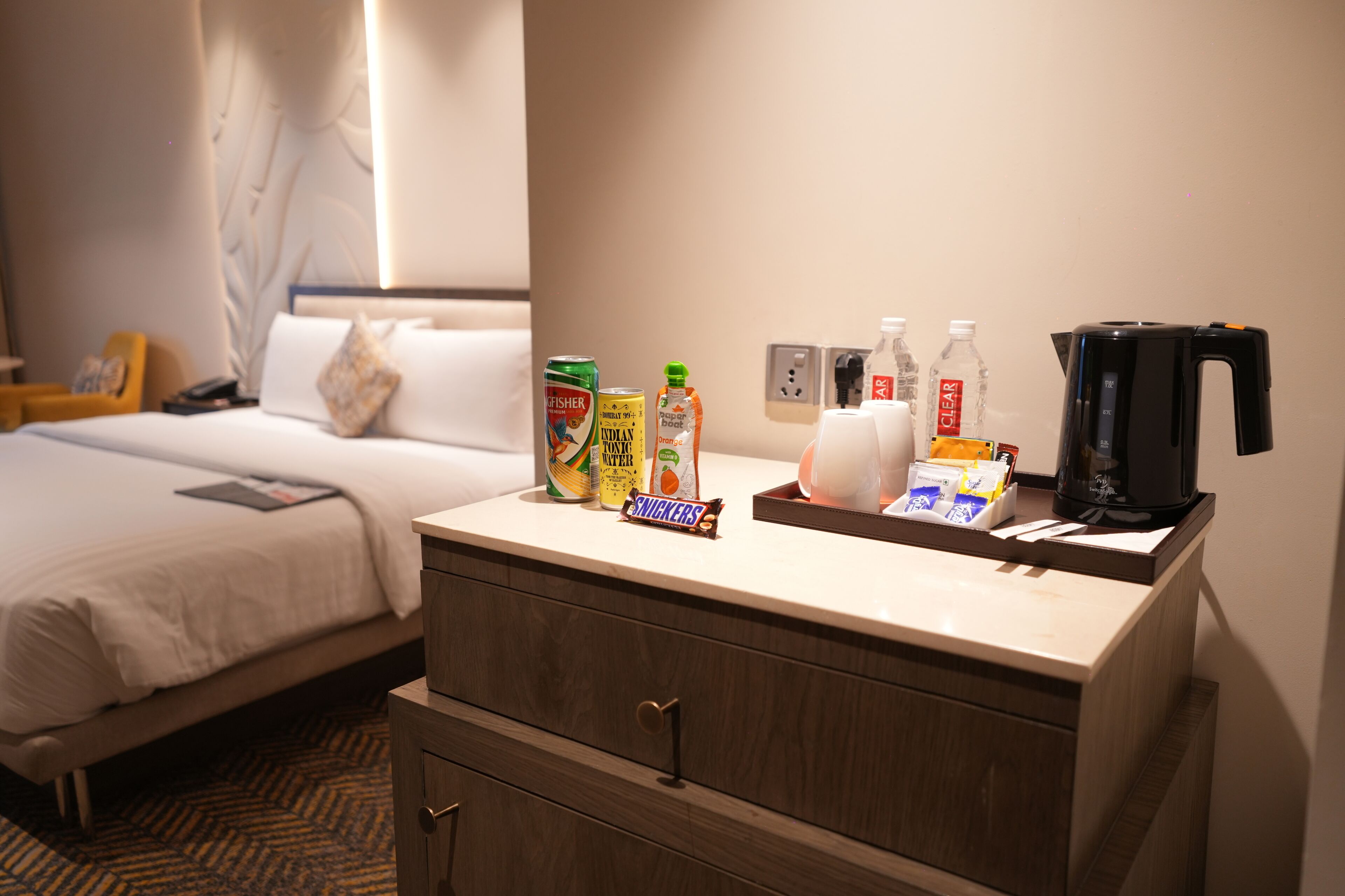 A hotel room with a bed, a bedside table with snacks, a coffee maker, and a wall socket.