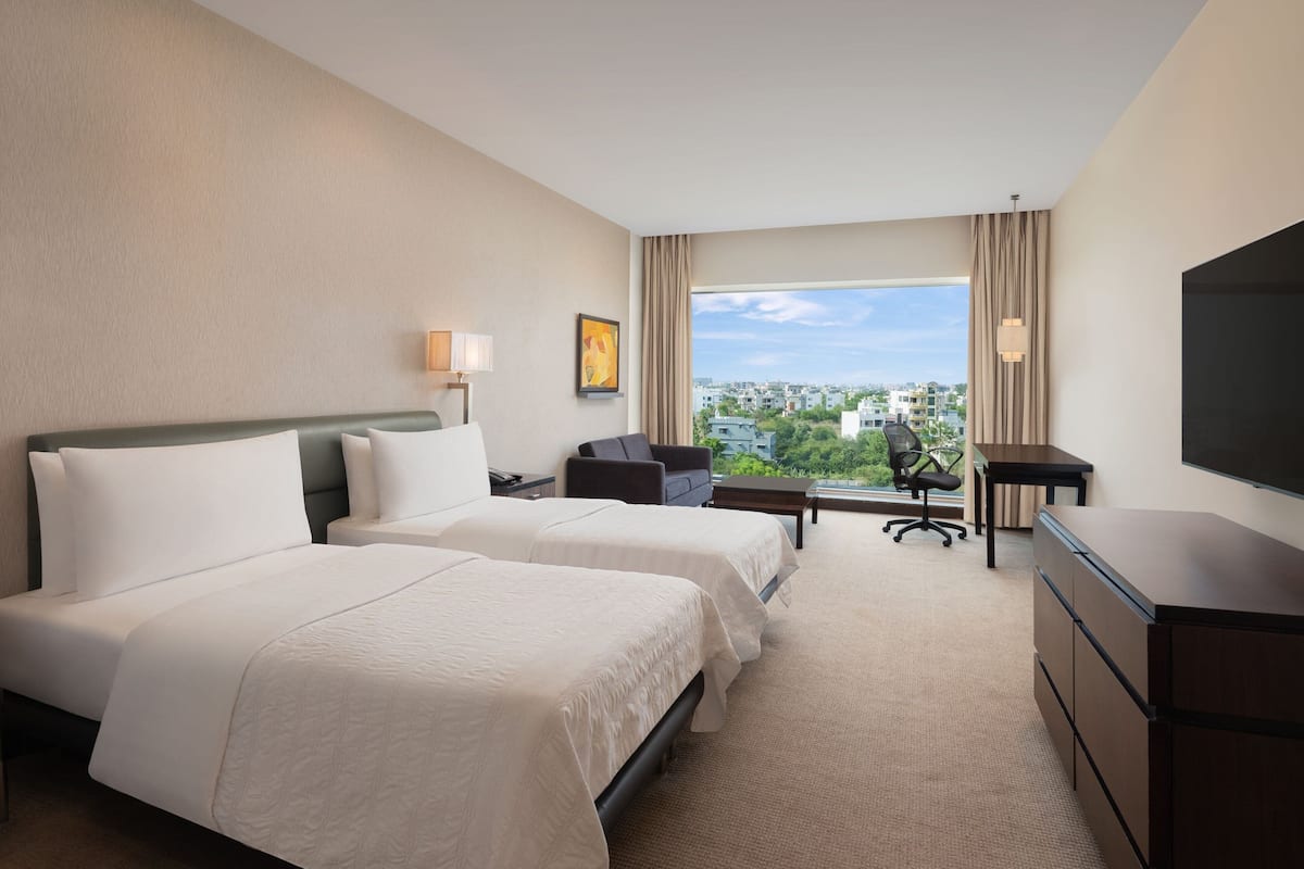 A hotel room with two beds, a desk, a chair, a TV, and a view of the city.