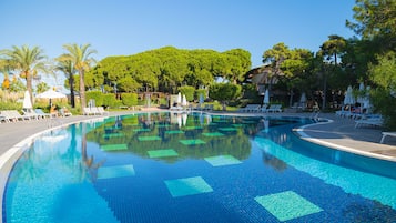 A large, clear swimming pool with tiled patterns, surrounded by lounge chairs and umbrellas, with trees and a clear sky in the background.