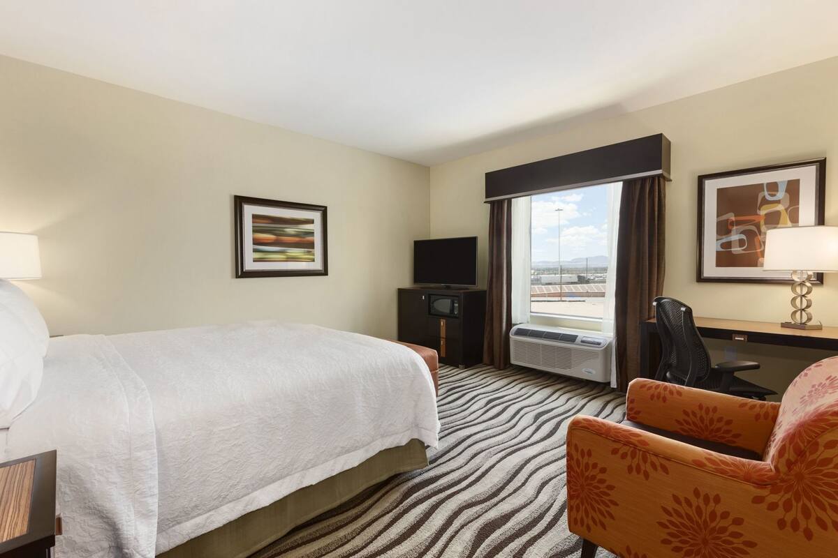 A hotel room with a bed, a desk with a chair, a television, and a window with curtains.