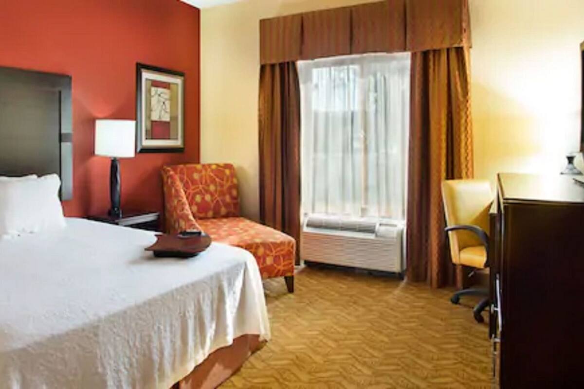 A hotel room with a large bed, a desk, a chair, a window with curtains, and a framed picture on the wall.