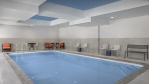 Una piscina interior con agua cristalina, rodeada de sillas y mesas blancas, y un banco.