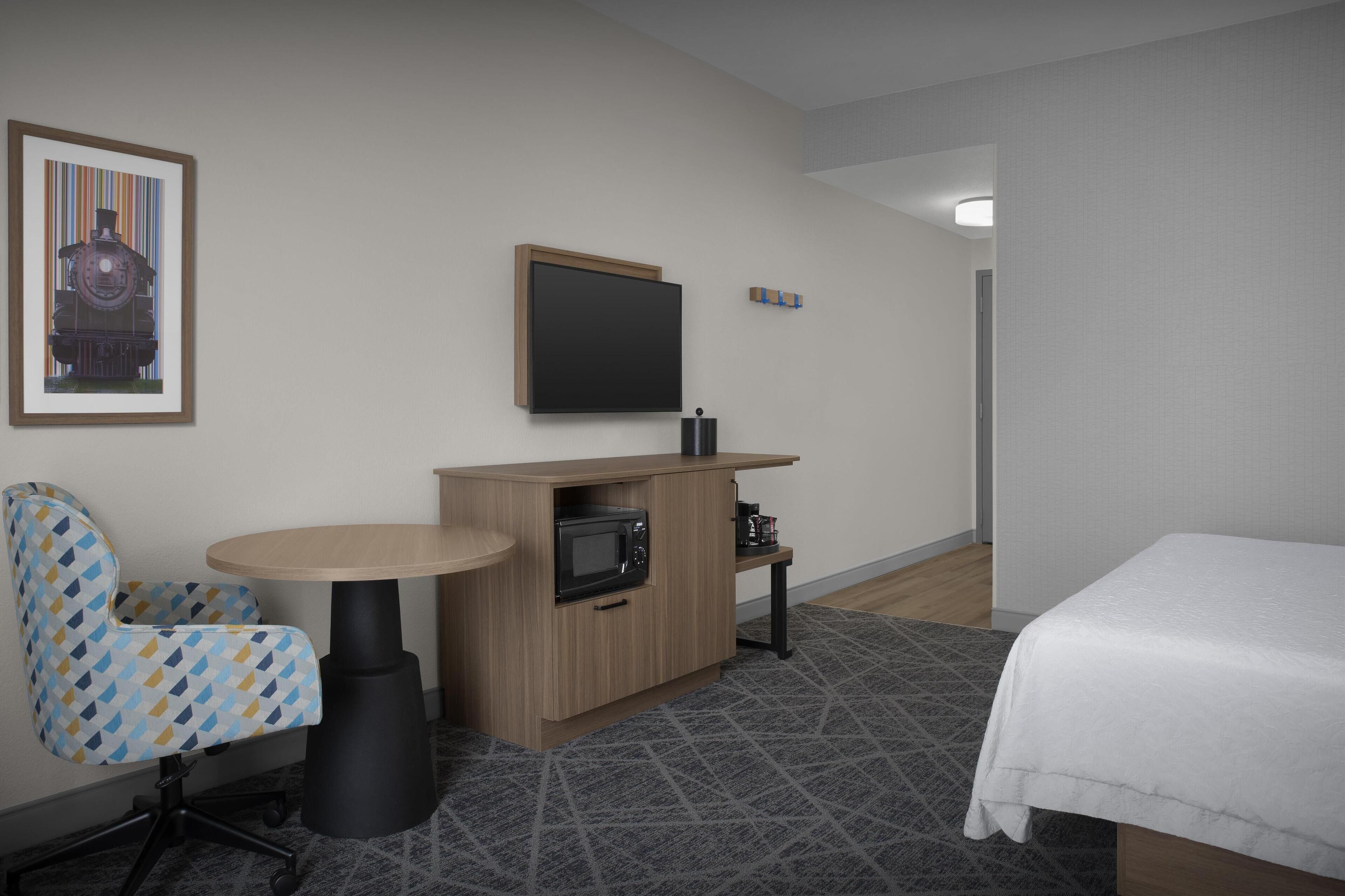 Una habitación de hotel con cama, escritorio con microondas, silla, televisión y un cuadro en la pared.