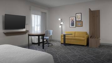 Habitación de hotel con cama, escritorio, sofá amarillo, televisor, lámpara y cuadros en la pared.