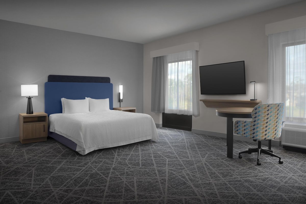 A hotel room with a bed, a desk, a chair, a TV, and a window with curtains.