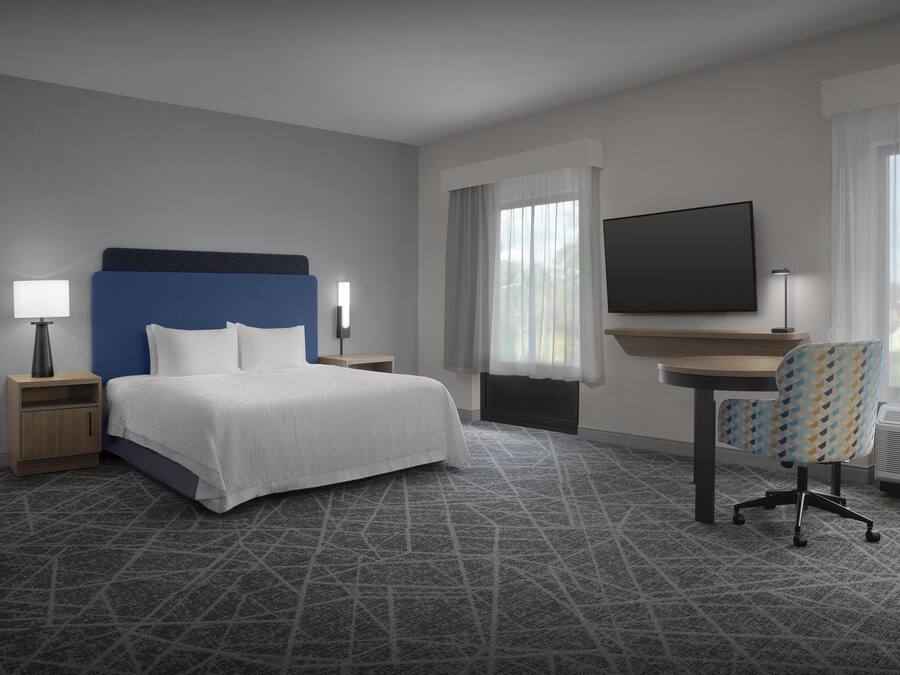 Una habitación de hotel con cama, escritorio, silla, televisor y ventana con cortinas.