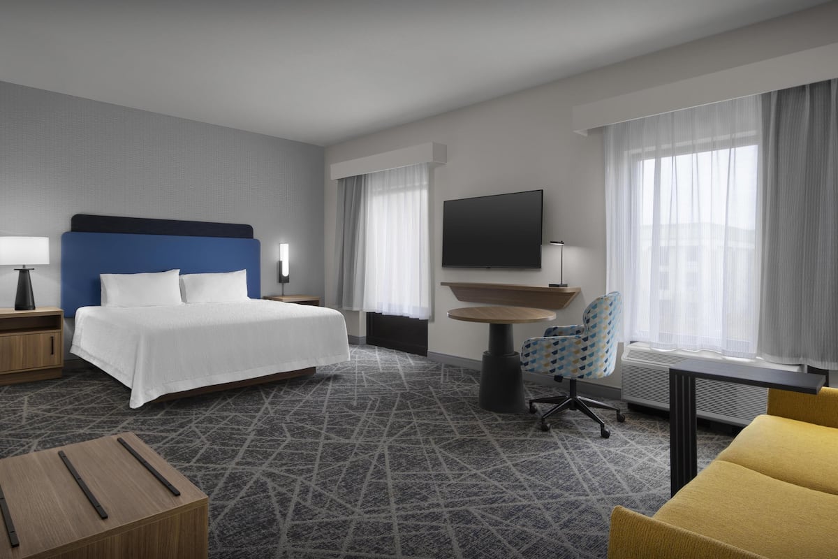 A modern hotel room with a large bed, a desk, a chair, a TV, and a window with curtains.