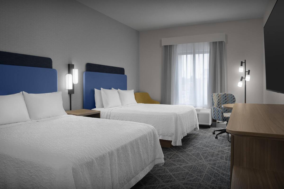 A hotel room with two beds, a desk, a chair, and a lamp.