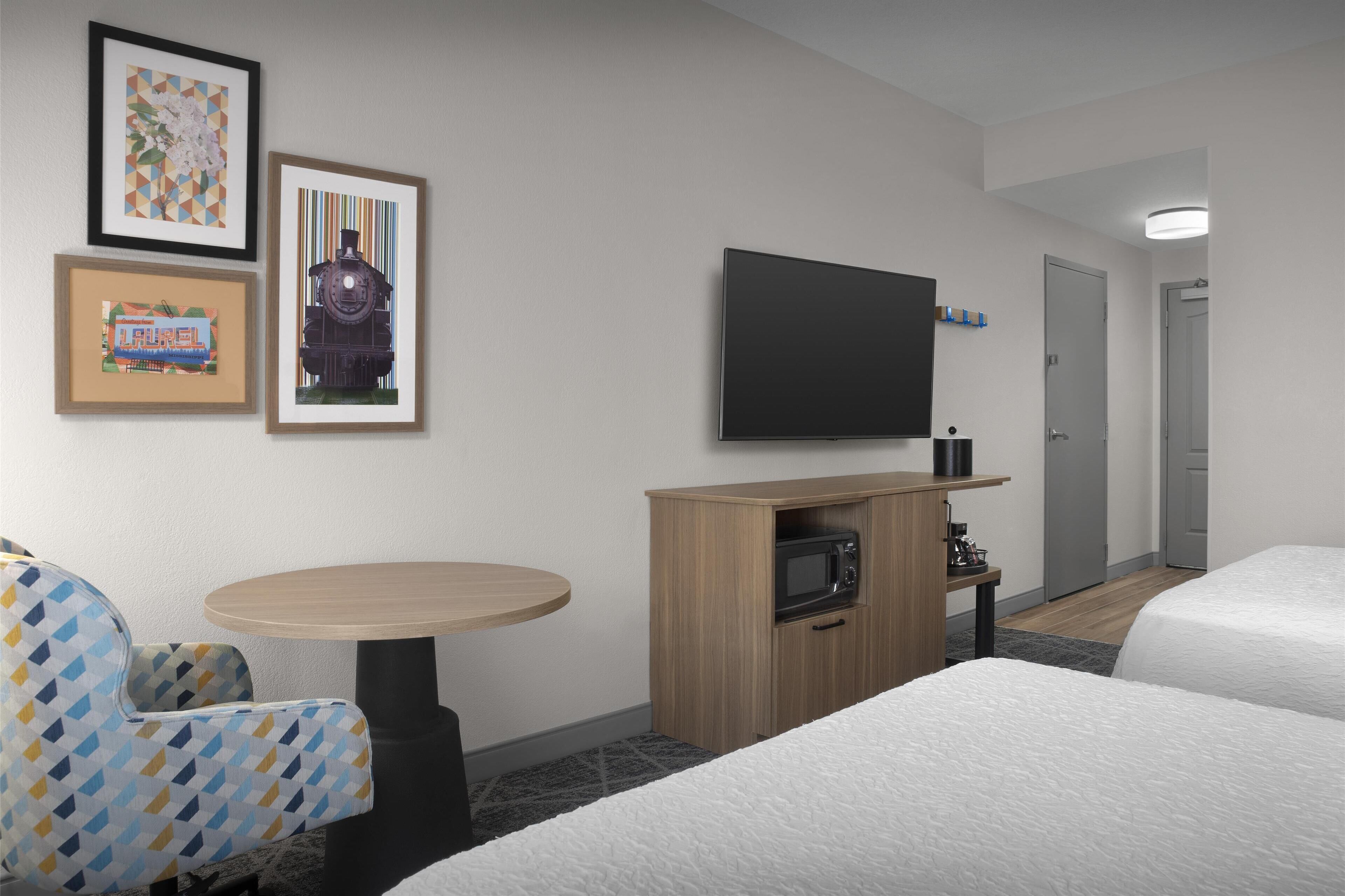 Habitación de hotel con dos camas, un televisor montado en la pared, un pequeño mueble de madera con microondas, una mesa redonda y una silla con estampado geométrico.
