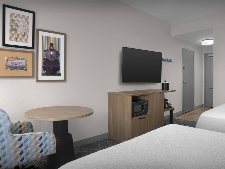 Habitación de hotel con dos camas, un televisor montado en la pared, un pequeño mueble de madera con microondas, una mesa redonda y una silla con estampado geométrico.