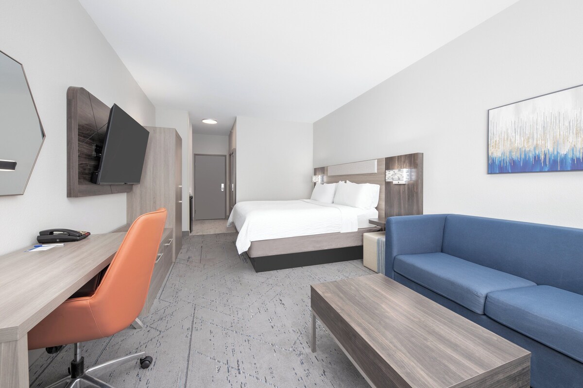 A modern hotel room with a bed, a desk with a chair, a sofa, a TV mounted on the wall, and a painting.