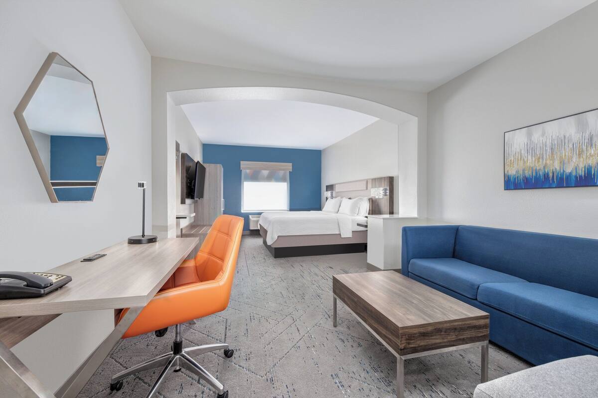A modern hotel room with a desk, orange office chair, blue sofa, and a bed with a white headboard.