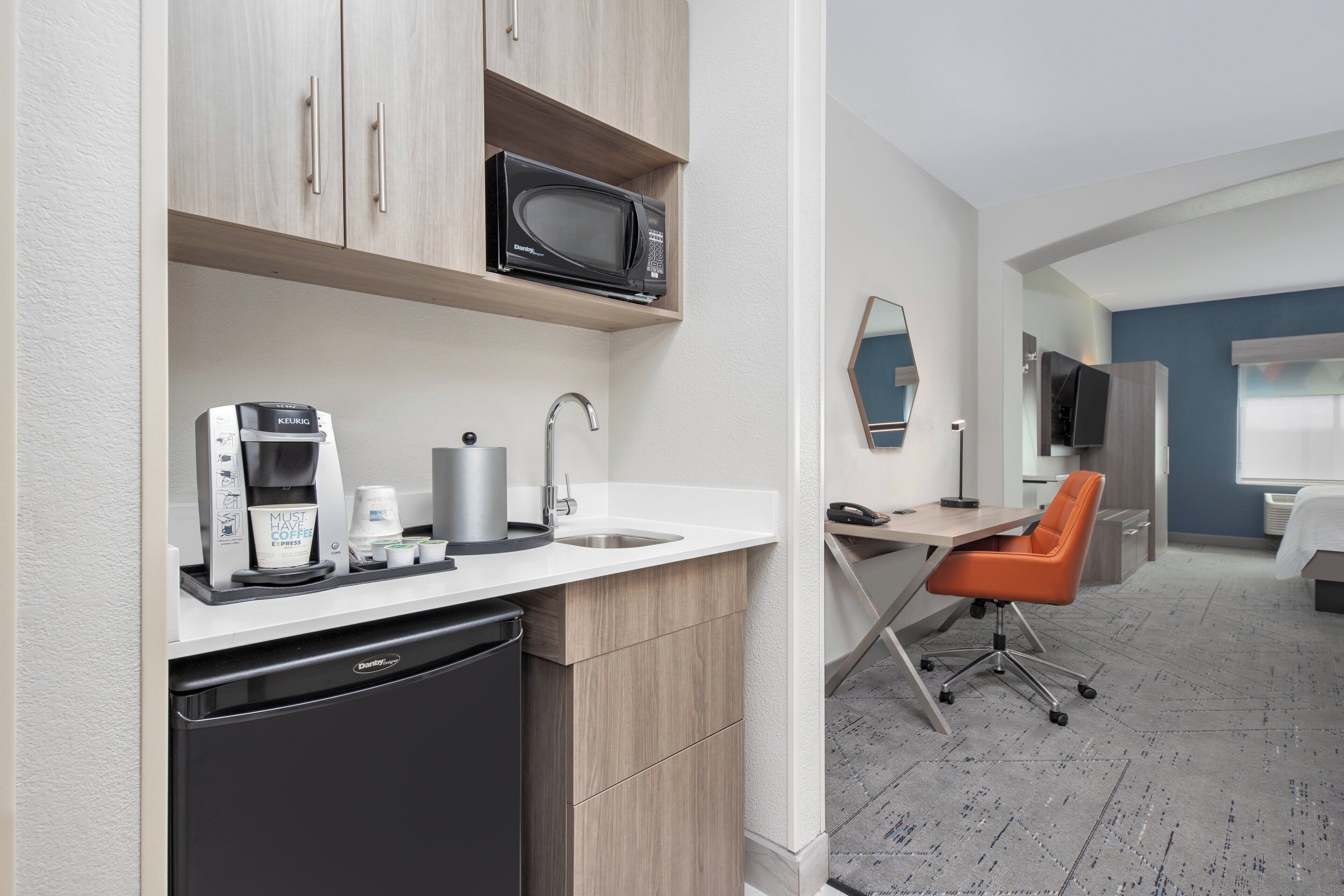 Una kitchenette moderna con microondas, cafetera y lavabo. Incluye un lavavajillas compacto y una pequeña zona de trabajo con escritorio y silla.