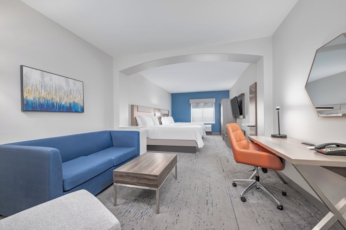 A hotel room with a blue sofa, a wooden coffee table, an orange office chair, a desk with a phone, and a bed with white bedding.