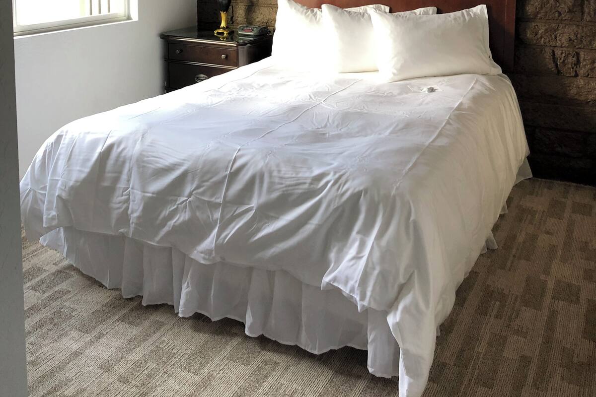 A bedroom with a brick wall, a wooden headboard, a white bedspread, and a nightstand with a lamp.