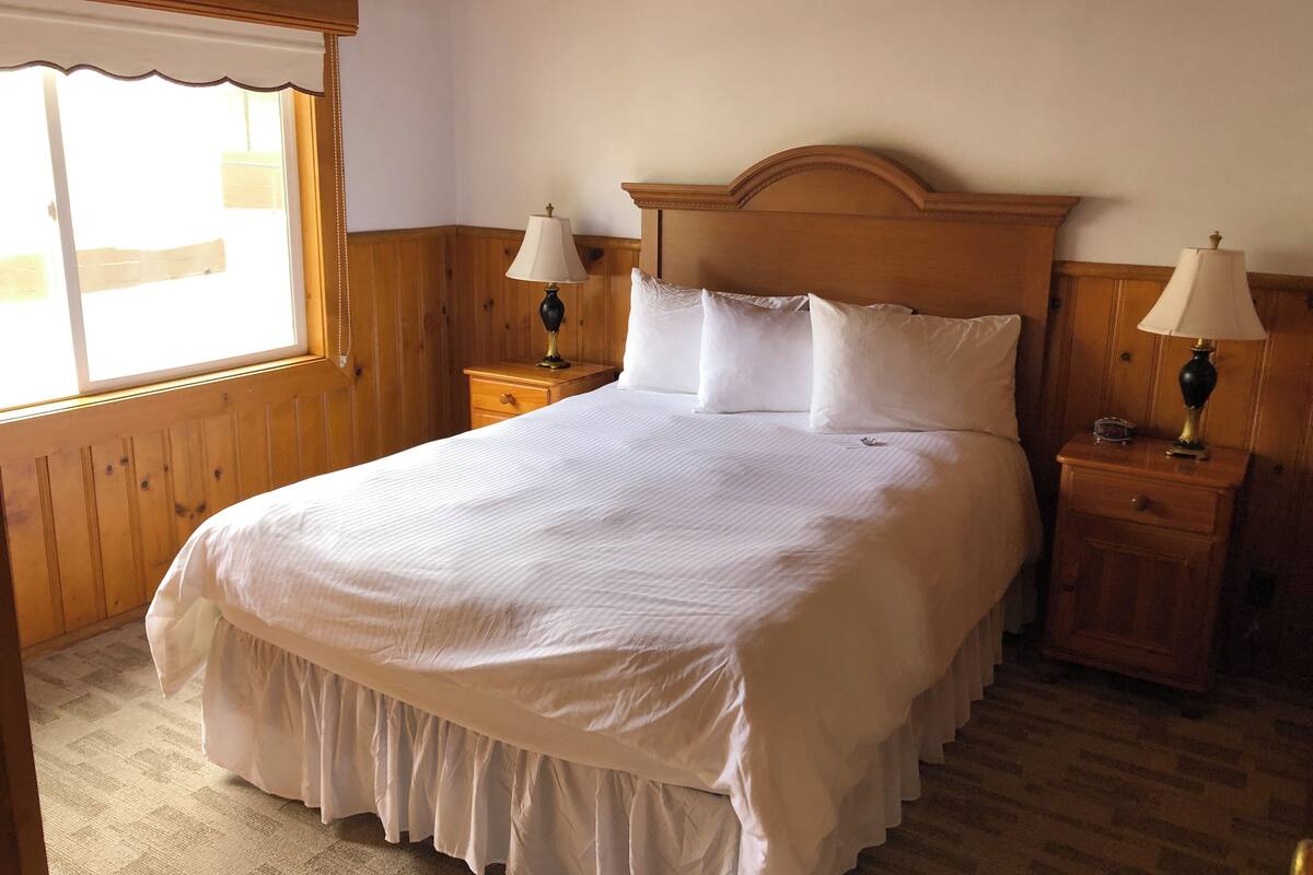 A neatly made bed with a wooden headboard, two bedside tables with lamps, and a window with a white valance.